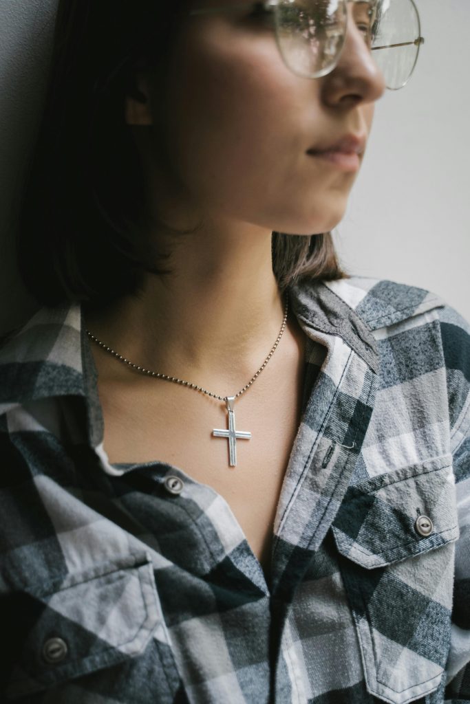 A close-up of a woman in glasses wearing a cross pendant necklace and plaid shirt indoors.